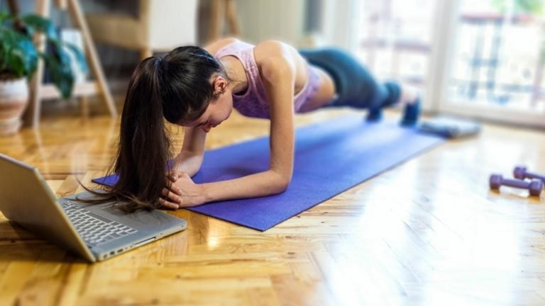 Uma mulher fazendo o exercício prancha, sobre um tapete lilás de frente para um laptop